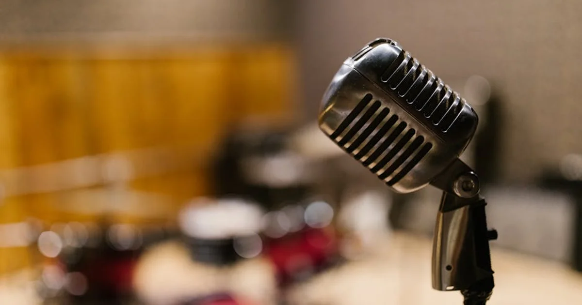 A professional Nigerian male voice actor in his home studio, speaking into a large condenser microphone, wearing headphones, with acoustic foam panels visible on the wall behind him. The image conveys professionalism and high-quality remote recording.